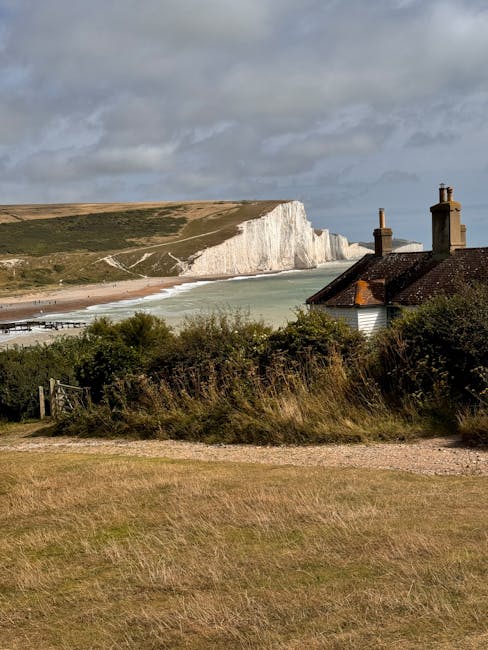 A scenic landscape of white chalk cliffs with rugged, vertical faces and grassy top edges, set against a partly cloudy sky. The cliffs show layers of white chalk with some areas covered in patches of 