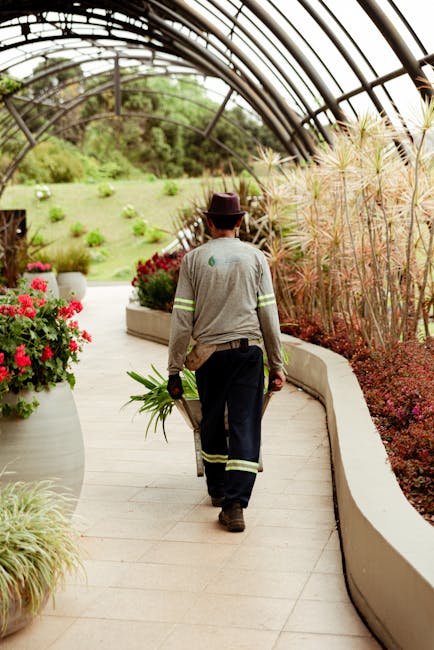 A gardener, dressed in a grey uniform with reflective strips on the sleeves and leg cuffs, is walking along a curved, light-colored paved pathway inside a large glass greenhouse. The gardener wears a
