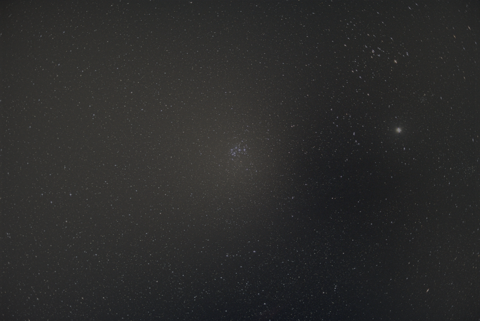 A night sky filled with numerous small, bright stars scattered across the dark expanse, with a faint cluster of stars visible towards the upper left and a brighter, more prominent star or planet towar